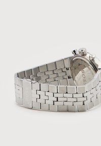 Ασημένιο μεταλλικό λουράκι ρολογιού με κούμπωμα, μερικώς ανοιχτό, προσαρτημένο σε στρογγυλό καντράν χρονογράφου που δείχνει το όνομα της μάρκας στην πίσω πλευρά.