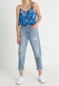 Donna che indossa un top senza maniche blu a fiori, jeans chiari strappati e arrotolati, e sneakers bianche, in piedi con una mano in tasca.