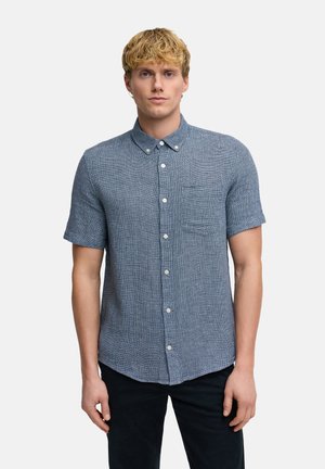 Nuori mies, jolla on vaaleat hiukset, pukeutuneena lyhythihaiseen siniseen kuvioituun kauluspaitaan ja mustiin housuihin, seisoo yksiväristä vaaleaa taustaa vasten.