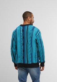 Maglione lavorato a maglia con texture in tonalità di blu e turchese, caratterizzato da motivi intricati. Rifinito con orlo e polsini a coste neri.