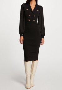 Robe midi noire côtelée avec un design à double boutonnage, boutons dorés et manches bouffantes, associée à des bottes à talons aiguilles crème arrivant au genou.