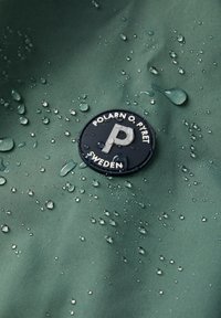 Waterafstotende groene stof met waterdruppels, met een cirkelvormig zwart logo met witte tekst en een opvallende letter "P."