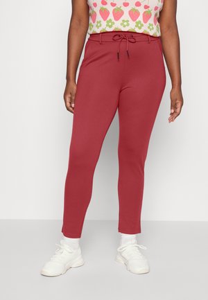 Kobieta ubrana w czerwone, wysokowciskowe, dopasowane spodnie z szlufkami, zawiązane z przodu, zestawione z białymi trampkami i bluzką w truskawkowy wzór.