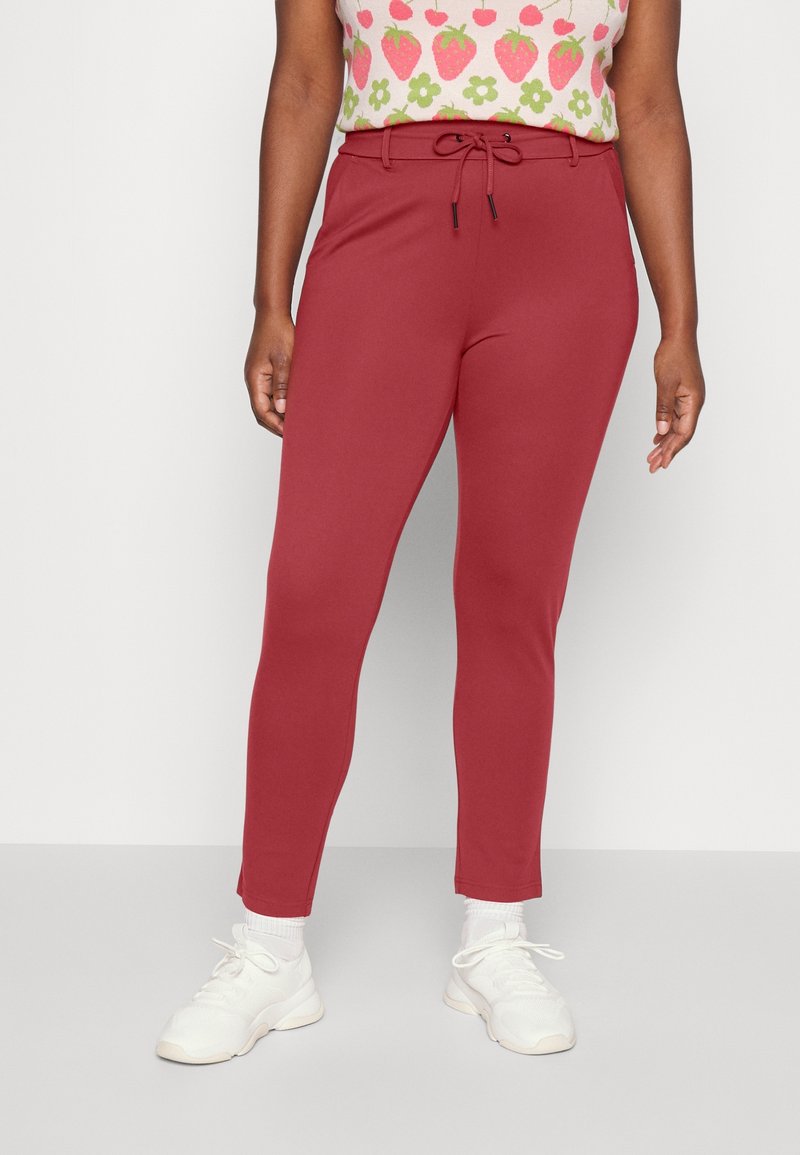 Femme portant un pantalon rouge taille haute et ajusté avec passants pour ceinture, noué à l'avant, assorti à des baskets blanches et un haut à motif fraises.