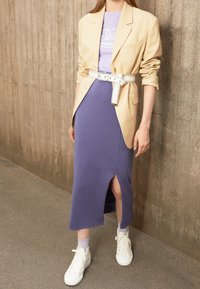 Béžový blazer přes levandulové šaty s bočním rozparkem, stáhnuté bílým páskem. Bílé vysoké tenisky dokončují outfit.