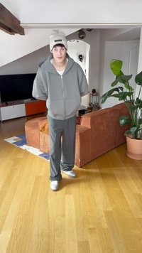 Grå zip-up hoodie och matchande byxor, bärs med en beige mössa och vita sneakers, i en modern inomhusmiljö med växter.