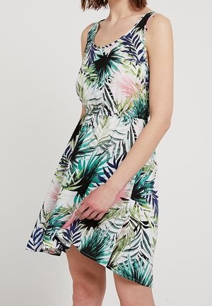 Femme portant une robe blanche sans manches avec des motifs de feuilles tropicales vertes, bleues, roses et noires, tenant l'ourlet de la jupe avec sa main droite.