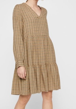 Kobieta w luźnej, długiej, warstwowej sukience w beżowo-brązową kratę z dekoltem w kształcie litery V, stojąca na tle jasnego, jednolitego tła.