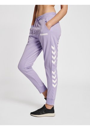 Personne portant un pantalon de survêtement lilas Hummel avec des bandes chevron blanches et des baskets noires, debout les mains dans les poches sur un fond blanc.