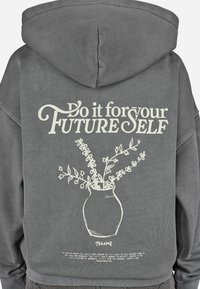 Γκρι υπερμεγέθη φούτερ με κουκούλα, που διαθέτει κεντραρισμένο λευκό γραφικό με μια βάση και φυτά, καθώς και κείμενο που λέει "Κάντο για τον ΜΕΛΛΟΝΤΙΚΟ ΕΑΥΤΟ σου." Υφή υφάσματος.