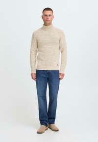 Hombre vistiendo un suéter de cuello alto beige, jeans azules y zapatos beige, de pie mirando hacia adelante contra un fondo blanco liso.