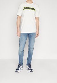Vit kortärmad t-shirt med en svart och neongrön grafisk design, tillsammans med ljusblå slim-fit jeans och vita sneakers med marinblå detaljer.
