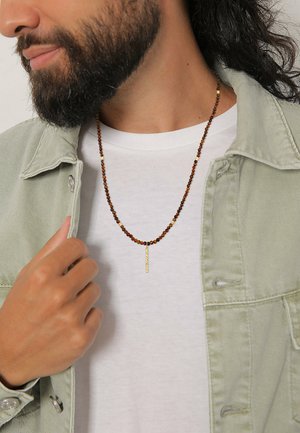 Brown beaded necklace with gold accents, featuring a vertical bar pendant. Worn over a white shirt and paired with a light green denim jacket.