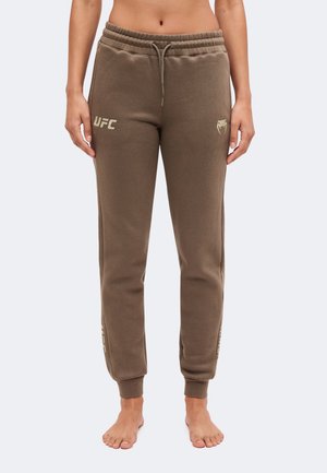 Persona descalza que lleva pantalones jogger marrones con la marca UFC, puños elásticos y cintura con cordón, de pie frente a un fondo liso.