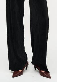 Άτομο που φοράει μαύρο πλισέ παντελόνι με φαρδιά πόδια και σκούρα καφέ γόβες με ανοιχτή φτέρνα και μυτερή μύτη, στέκεται σε λευκή επιφάνεια.