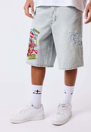 Pantalones cortos de mezclilla gris claro con parches coloridos de la Ruta 66 y gasolina, usados con zapatillas blancas y calcetines blancos con logo.