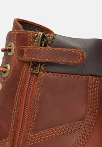 Bota de piel marrón con cierre de cremallera, costuras en contraste y un upper texturizado con un acabado marrón suave cerca de la apertura.