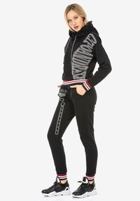 Schwarzer Hoodie mit reflektierendem Text, rot-weißen gestreiften Bündchen, passenden schwarzen Jogginghosen mit silberner Kettenmuster und Taschen.