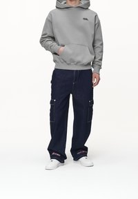 Szara bluza z kapturem z przednią kieszenią i logo, zestawiona z luźnymi ciemnymi dżinsowymi spodniami typu cargo z bocznymi kieszeniami i białymi akcentami, noszona z białymi trampkami.