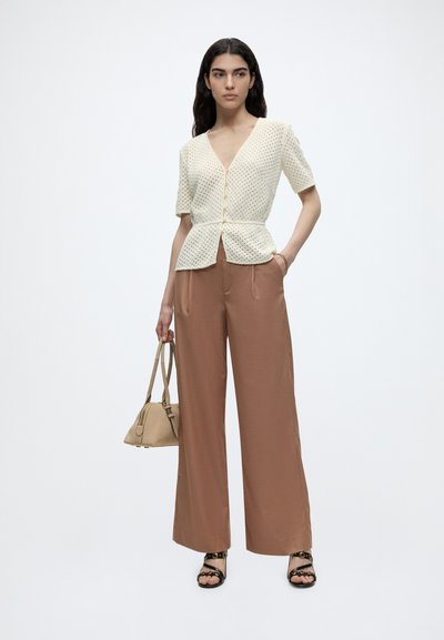 Γυναίκα που φοράει μπεζ πλεκτό τοπ με κοντά μανίκια, καφέ παντελόνια με φαρδιά μπατζάκια, μαύρα σανδάλια και κρατά μια μπεζ τσάντα με το ένα χέρι στην τσέπη.