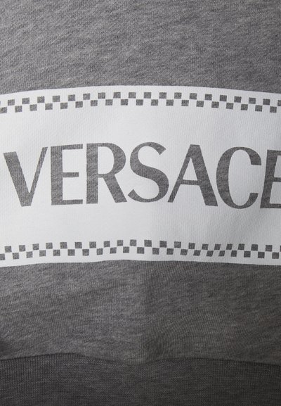 Tessuto grigio con banda bianca che mostra "VERSACE" in caratteri grandi, con accenti a scacchi. Texture morbida, design informale.