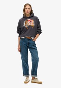 Haut à capuche gris foncé avec un graphique de tigre, associé à un jean bleu clair retroussé et des baskets beiges. Look décontracté avec une silhouette en couches.