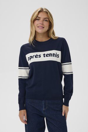 Marineblauer Strickpullover mit weißem Streifen und dem Text "après tennis". Gerippter Kragen und Bündchen. Weiche Textur, lässiges Design. Kombiniert mit dunklen Jeans.
