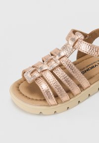 Sandale métallique rose doré avec plusieurs sangles horizontales et un semelle intérieure beige contournée. Semelle extérieure en caoutchouc avec un détail de grip texturé.