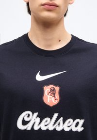 Άτομο που φοράει μπλε σκούρο μπλουζάκι Chelsea FC με λευκό λογότυπο Nike και κείμενο «Chelsea», κοντινή λήψη που δείχνει τον λαιμό και το πάνω μέρος του στήθους.