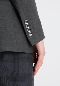 Manche d'un blazer gris avec quatre boutons noirs, posée à côté d'une main et associée à un pantalon à carreaux foncés.