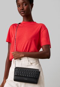 Svart kviltad crossbody-väska med en slät bottenpanel, silverkedjesnor och logotypdetalj, bärs mot en röd skjorta och ljusa byxor.
