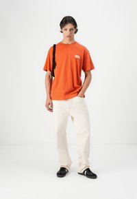 Oranje katoenen t-shirt met korte mouwen en een borstprint, gecombineerd met off-white relaxte broek en zwarte sneakers.