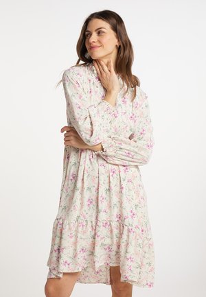 Donna con capelli castani che indossa un vestito bianco a maniche lunghe con stampa floreale rosa, in piedi con le braccia incrociate e guarda di lato.