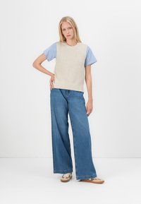 Gilet en tricot beige sur une chemise à rayures bleues et blanches, associé à un jean bleu à jambes larges et des chaussures à motifs, le tout sur fond blanc.