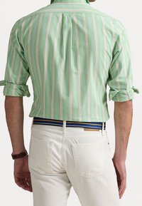 Camisa a rayas verdes con líneas en rosa claro y azul, con mangas enrolladas. Combinada con pantalones blancos y un cinturón de rayas multicolor.
