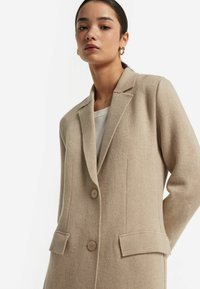 Beige Wollmischungsmantel mit Reverskragen, zwei Knöpfen und seitlichen Taschen; verfügt über eine taillierte Passform und einen strukturierten Stoff.