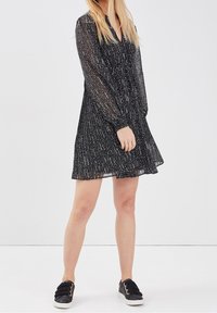 Robe noire transparente à manches longues, motif à pois, encolure boutonnée et jupe plissée. Associée à des baskets noires à enfiler.