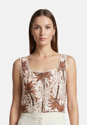 Donna con capelli castani lisci che indossa una canotta bianca senza maniche con motivi a foglia marroni e bottoni frontali, abbinata a pantaloni beige.