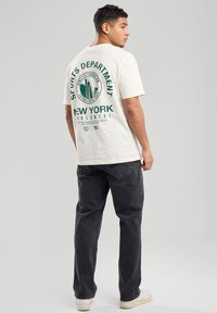 T-shirt crème avec un texte graphique vert et une silhouette de ville au dos, associé à un jean sombre et des baskets blanches, affichant une coupe classique.