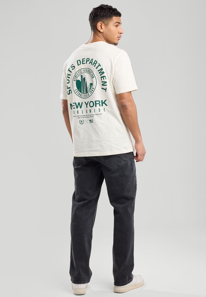 T-shirt crème avec un texte graphique vert et une silhouette de ville au dos, associé à un jean sombre et des baskets blanches, affichant une coupe classique.