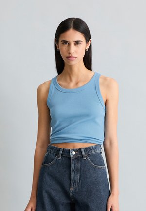 Jeune femme aux longs cheveux noirs portant un crop top bleu clair sans manches et un jean taille haute bleu foncé sur un fond gris uni.