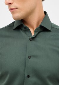 Camicia button-up verde scuro, con texture e colletto, caratterizzata da bottoni neri e un motivo sottile, esposta su uno sfondo neutro.