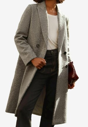 Vrouw draagt een gestructureerde grijze dubbelgeknoopte jas over een wit shirt en donkere broek, en houdt een kleine donkerrode clutch vast.