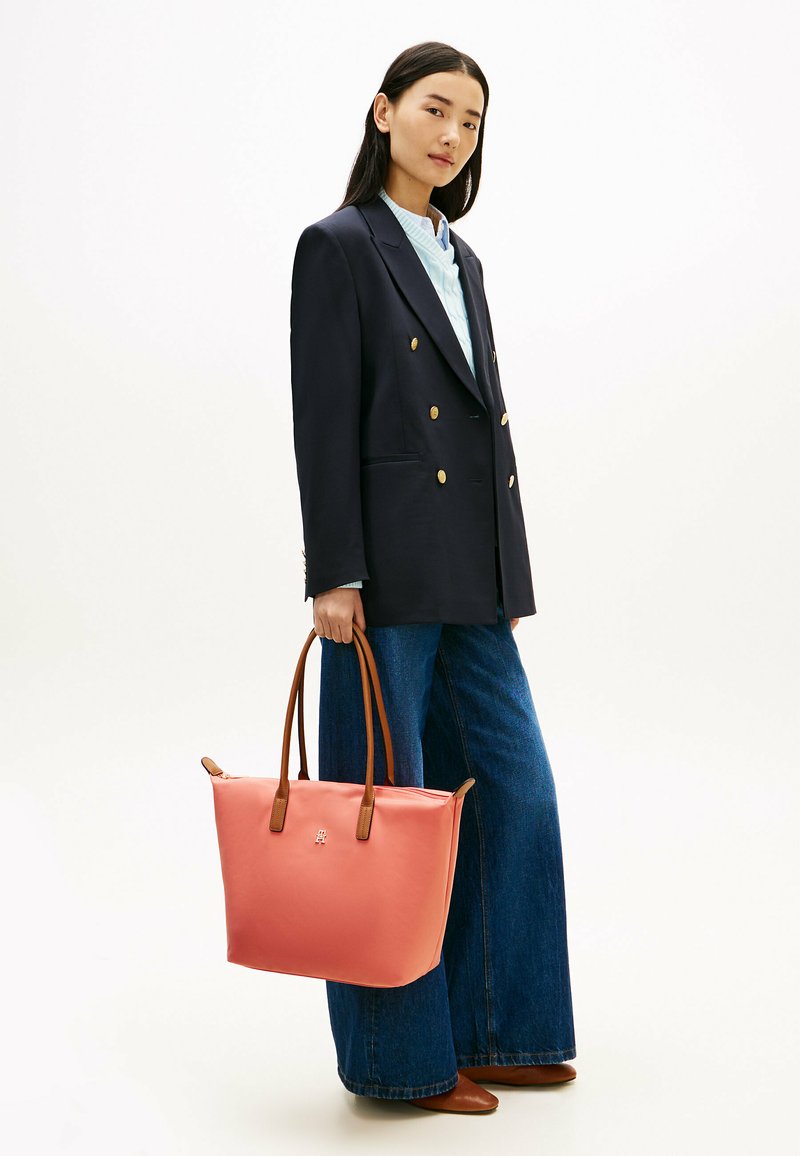 Femme portant un blazer bleu marine, un pull bleu clair, un jean à jambes larges, des chaussures marron, tenant un grand sac fourre-tout corail avec des poignées marron.