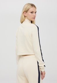 Skórzana bluzka w kolorze kremowym z granatowymi paskami po bokach, z wysokim kołnierzem i miękką teksturą. W komplecie z pasującymi kremowymi joggerami.