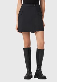 Zwarte mini-rok met twee voorzakken, een knoopsluiting en een gladde textuur. Gepaard met zwarte kniehoge laarzen.