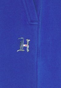 Blå pullover med et sølvbroderet "H"-logo på brystet, lavet af struktureret stof og med v-hals design.