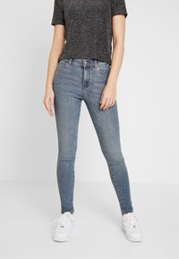 Smala blå denimjeans med en blekt finish, hög midja och klassisk femfickdesign, kombinerat med en svart prickig t-shirt och vita sneakers.