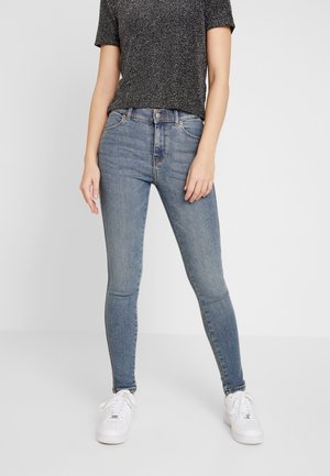 Jeans de denim azul ajustados con un acabado desgastado, cintura alta y diseño clásico de cinco bolsillos, combinados con una camiseta negra con manchas y zapatillas blancas.