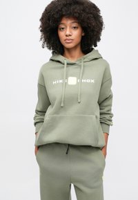 Γυναίκα με σγουρά μαλλιά που φοράει ελιάς πράσινο φούτερ Nike Shox με κουκούλα και ταιριαστό παντελόνι, στέκεται με τα χέρια στις τσέπες μπροστά σε ένα απλό φόντο.